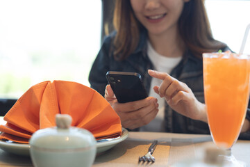 Happy young asian woman using mobile phone and drinking orange juice in restaurant, cafe interior. Smiling woman using smartphone for social media network, surfing internet, close up