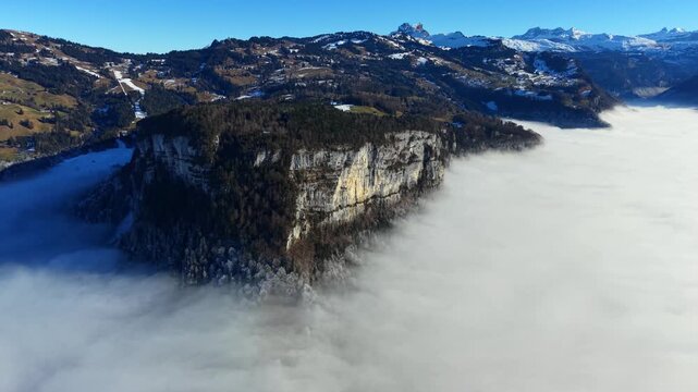 Dramatic drone shot of a massive limestone cliff face emerging from a thick blanket of clouds in Canton Schwyz Muotathal.