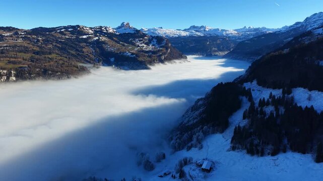 Beautiful drone establishing shot looking out over a thick layer of clouds, fog filling the valley below the snowy peaks of Stoos.