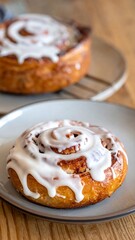 A close-up of a glazed cinnamon roll on a gray plate, with another roll on a rack in soft focus in the background