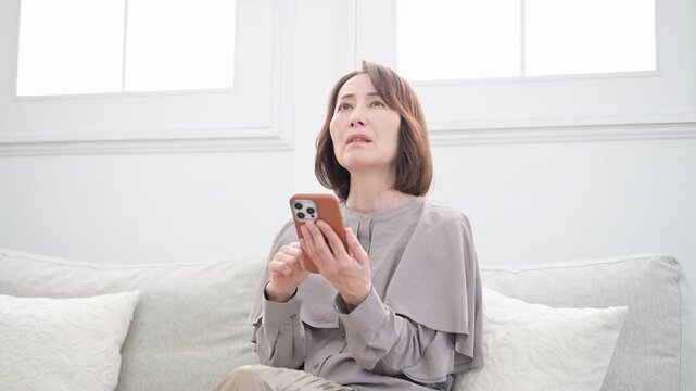ソファに座りスマホを操作して予約や問い合わせで困る、悩む考える年配女性の動画