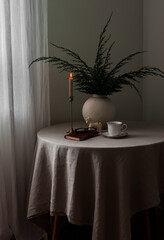 A round table with a gray tablecloth, a vase with juniper branches, a candle, and a cup of coffee in the living room