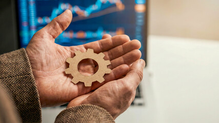 Business high level planning concept gear held in hand near computer screen showing financial chart metaphor for management and strategy focus