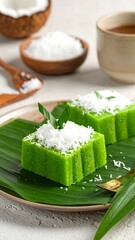 Two green steamed cakes with coconut topping on a banana leaf, with tea and coconut