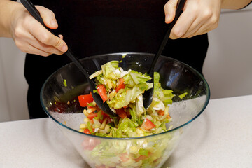 Person's hands using salad servers to mix a bowl of fresh vegetables and ingredients, preparing a nutritious meal