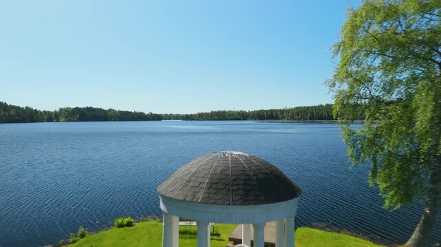 White gazebo on grassy island connected by wooden pier amid serene lake near H&aring;verud village Mellerud Sweden with birch trees blue sky