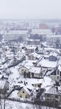Vertical Drone glide reveals a dense suburban grid of snow-covered private villas , framed by distant multi-story soviet era residential towers under heavy snowfall, Darzciems, Riga, Latvia.