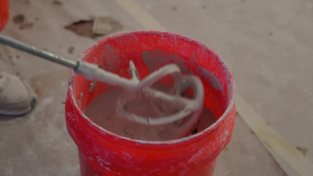 POV of worker mixing construction mortar in red bucket using a power mixer. Close up of renovation work, building preparation, and professional construction tools in use.