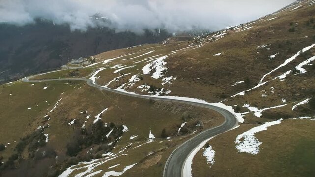 Cinematic aerial tilt-down revealing a winding mountain road near the French border in Larra-Belagua. Snow patches and vegetation dot the landscape under heavy winter clouds.