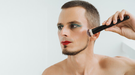 portrait of gay man with makeup and cosmetics on face. A handsome young transgender man with red lipstick on lips and painted eyes applies blush with a brush and does facial contouring