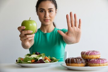  A woman holds a green apple in one hand while making a stop gesture toward a plate of donuts. Her focus is clearly on making a healthy dietary choice and resisting sugary temptations.