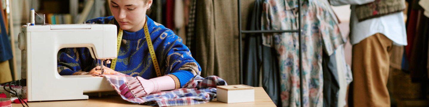 Young adult Caucasian woman using sewing machine repairing second hand clothing in workshop, measuring tape around neck, focused on garment, racks of clothes in background
