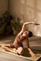 Obraz premium Caucasian young adult woman sitting on yoga mat performing seated side stretch with arm raised, wearing glasses, practicing yoga or pilates in bright indoor space