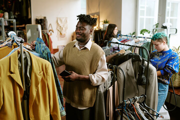 Fototapeta premium Young Black man browsing clothing racks in second hand store, holding smartphone, standing among diverse group of shoppers including young Caucasian woman with green hair searching for used clothes
