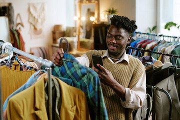 Young adult Black man browsing second hand clothing rack, holding smartphone and examining plaid...