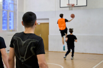 Indoor basketball action with a player jumping toward the hoop while others run and observe, capturing energy, teamwork, and training in a dynamic sports environment.