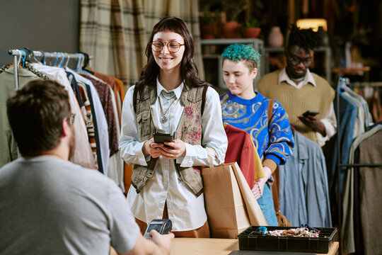 Young biracial woman smiling while holding smartphone and shopping bag, standing in line with young multiethnic adults at second hand clothing store checkout counter