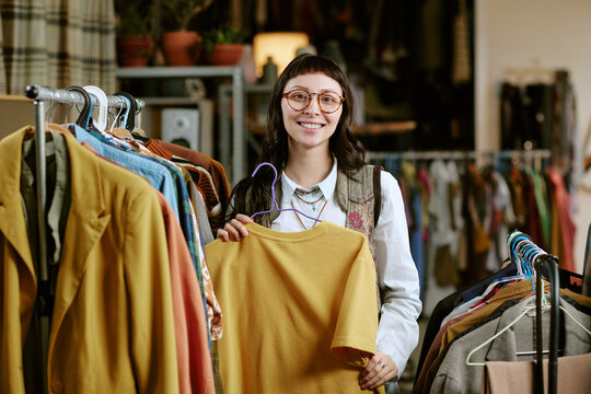 Portrait of young adult biracial woman smiling while holding second hand shirt on hanger in thrift store, surrounded by racks of used clothing, engaging in sustainable fashion shopping