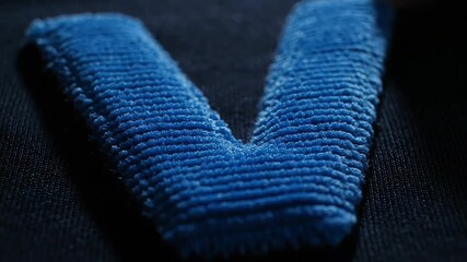 A close-up shot of a blue fabric letter V on a dark background, showcasing texture and detail from a macro viewpoint like sports scenes