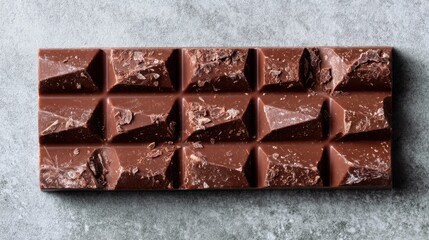 A chocolate bar with a distinct pattern sits on a gray surface. The bar shows signs of being opened and it waits to be enjoyed. The setting is simple and focuses on the chocolate.