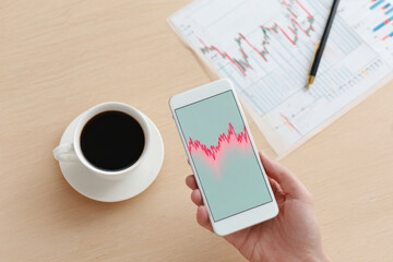 Close-up of hands with smartphone showing stock graph near cup of coffee. Concept of investment, market analysis, and financial management in warm light.