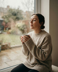 Woman in cozy knit sweater holding steaming cup of coffee by window on a rainy day enjoying the aroma