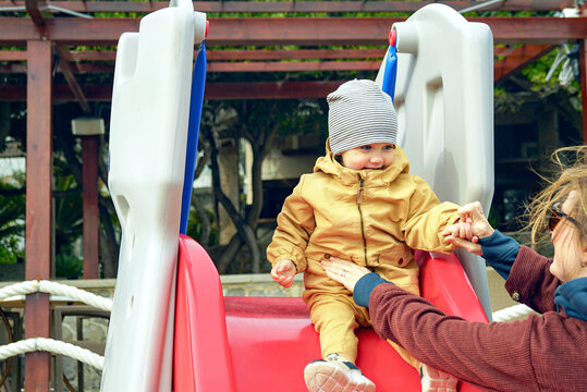 Joyful toddler sliding down colorful playground with mother's supportive hand nearby. Represents secure attachment parenting, supervised outdoor exploration, and child-centered recreation approach.