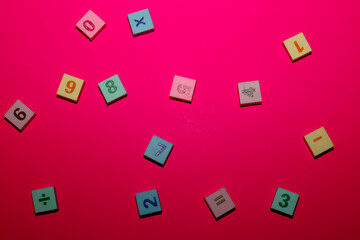 Colorful square tiles with numbers and mathematical signs are haphazardly scattered on a bright pink background. 
