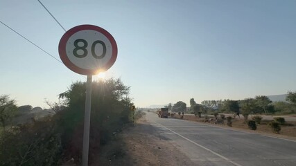 Tracking shot along rural highway passing a circular 80 speed limit road sign as trucks drive ahead, dry landscape and trees lining the road under low sun