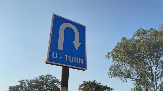 Low-angle orbiting shot around blue U-turn road sign with curved arrow symbol, set against clear sky and roadside trees, sunlight creating soft highlights on sign