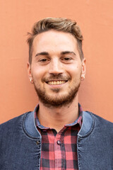 Obraz premium Smiling young man standing in front of a wall wearing a checkered shirt and denim jacket. He looks confident, friendly and approachable. Millennial generation and modern casual style concept.