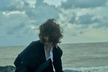 Fototapeta premium Windy coastal scene with an individual in a dark jacket by the ocean, as waves crash and clouds roll over the horizon, creating a moody seascape