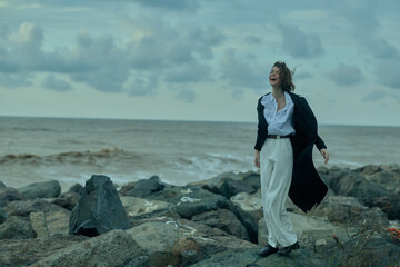 Woman on a rocky shoreline wearing a flowing coat, white pants and a crisp shirt, by the sea in...