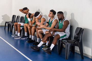 Diverse male basketball teammates sitting on chairs at gym wearing green and white uniforms