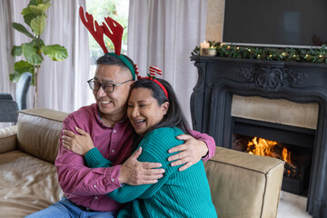 Naklejka premium Senior couple hugging while sitting on couch in living room with lit fireplace, antler headbands
