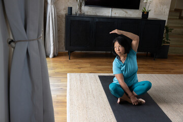Senior Asian woman practicing seated side stretch on yoga mat over woven rug in living room