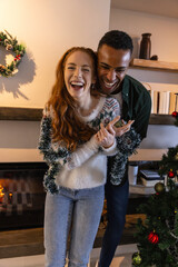 Naklejka premium Diverse couple embracing and decorating in living room with tinsel, garland and wreath