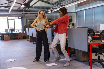 Diverse coworkers dancing playfully in business attire at open plan office among desks and laptops