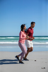 Naklejka premium Diverse couple jogging along sandy beach shoreline in athletic clothing and running shoes