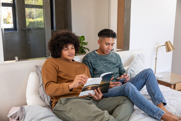Diverse male friends sitting on bed reading open book and scrolling smartphone in modern bedroom
