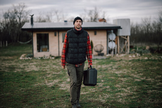 Man walks with a black case near a small cabin in the countryside