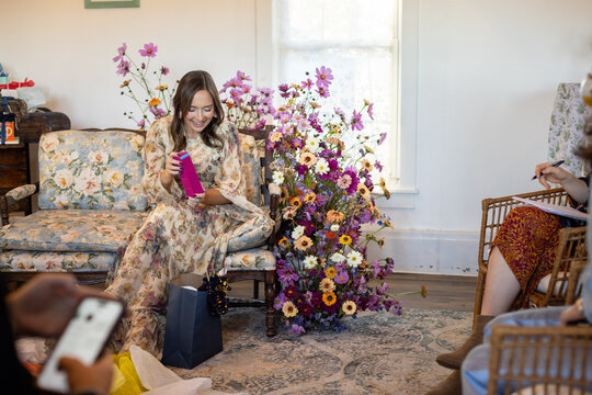 Woman on floral couch unpacks baby shower gifts surrounded by fl