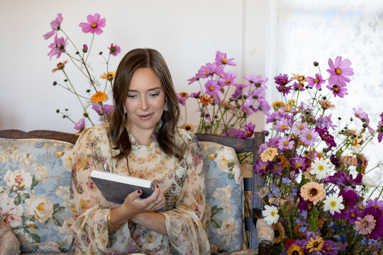Woman on couch surrounded by flowers looks at a book