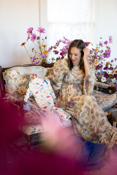 Woman on floral couch unpacks baby shower gifts surrounded by fl