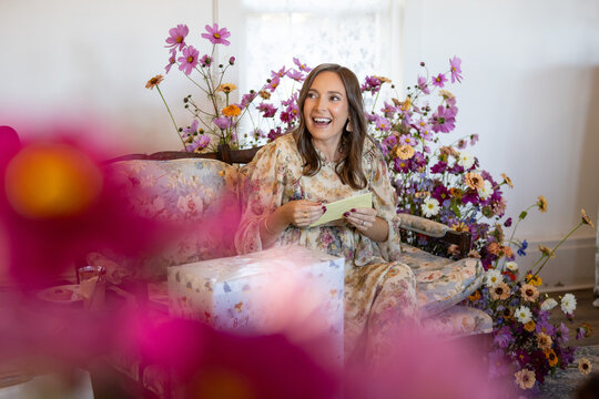 Woman in floral setting opens a card and laughs seated next to a