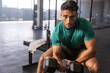 Man sitting on workout bench in gym holding hexagonal dumbbell near towel, water bottle
