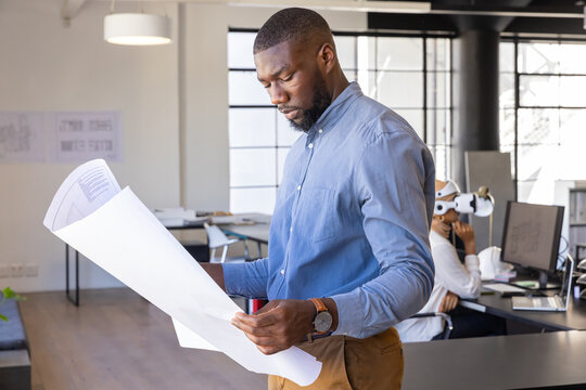 Diverse coworkers holding blueprints, using vr headset in modern open-plan office workspace - Powered by Adobe
