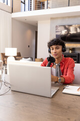 Naklejka premium Male broadcaster wearing headphones sitting at wooden desk in living room using laptop
