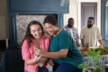 Obraz premium Diverse friends leaning over smartphone at kitchen island while slicing fruit and holding juice