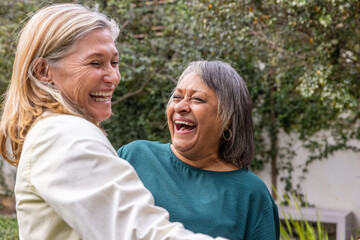 Diverse senior female friends embracing and laughing in garden wearing jacket and blouse